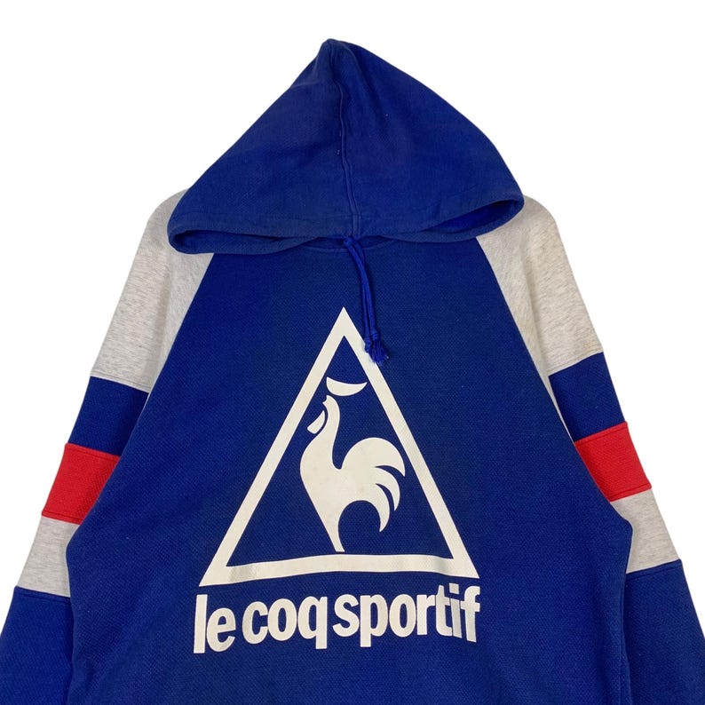 Puede incluir: Una sudadera con capucha azul, gris y roja con un logotipo de gallo blanco y el texto "le coq sportif" en la parte delantera.