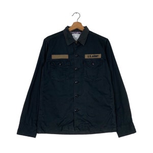 Peut inclure: Une chemise boutonnée à manches longues noire avec un patch marron qui dit "U.S. ARMY" sur la poitrine droite. La chemise a deux poches sur le devant et un col boutonné.