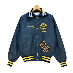Puede incluir: Una chaqueta de béisbol de satén azul marino con rayas amarillas y blancas en el cuello y los puños. La chaqueta tiene cierre a presión y presenta el texto "Magurasky" y "Glenbrook South Titans" con un gráfico de baloncesto en el pecho izquierdo. El número "93" está bordado en el lado derecho de la chaqueta.