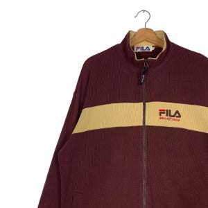 May include: A maroon zip-up jacket with a cream-colored horizontal stripe across the chest. The jacket features the "FILA" logo on the chest and collar. The jacket is hanging on a metal hanger.