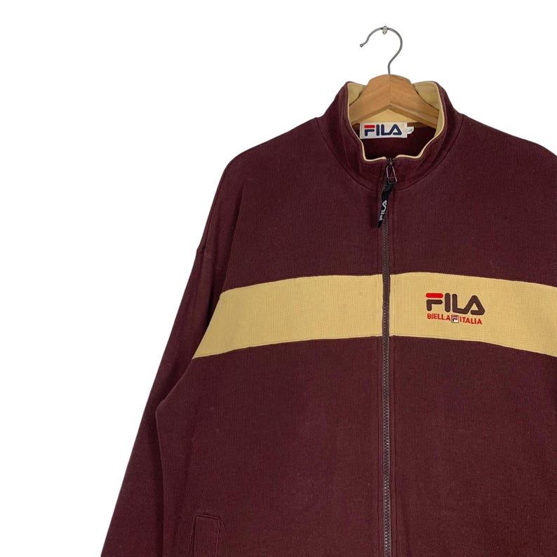 May include: A maroon zip-up jacket with a cream-colored horizontal stripe across the chest. The jacket features the "FILA" logo on the chest and collar. The jacket is hanging on a metal hanger.