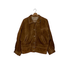 May include: A brown suede jacket with a button-up front, two front pockets, and a collared neckline. The jacket is hanging on a wooden hanger against a white background. The jacket has a classic, vintage style.