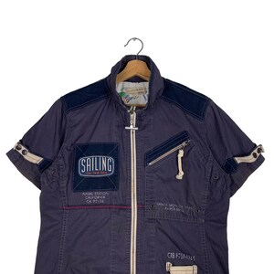 May include: A navy blue short-sleeved jacket with a zip fastening. The jacket has several pockets and features a patch with the text "SAILING ON THE SEA". Other text on the jacket includes "NAVAL STATION CALIFORNIA CA 92136", "SANTA MON.CA 1900 AVIREX ME", and "CRB P-75A-1965".