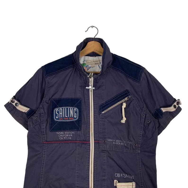 May include: A navy blue short-sleeved jacket with a zip fastening. The jacket has several pockets and features a patch with the text "SAILING ON THE SEA". Other text on the jacket includes "NAVAL STATION CALIFORNIA CA 92136", "SANTA MON.CA 1900 AVIREX ME", and "CRB P-75A-1965".