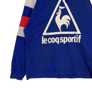 Puede incluir: Sudadera azul con un logotipo de gallo blanco en un tri&aacute;ngulo y el texto "le coq sportif" en blanco.