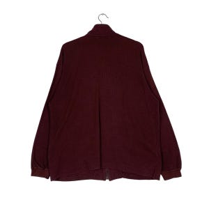May include: A burgundy-colored long-sleeve zip-up shirt hanging on a hanger. The shirt has a high collar and a full-length zipper. The sleeves have ribbed cuffs. The shirt is made of a soft, textured fabric.