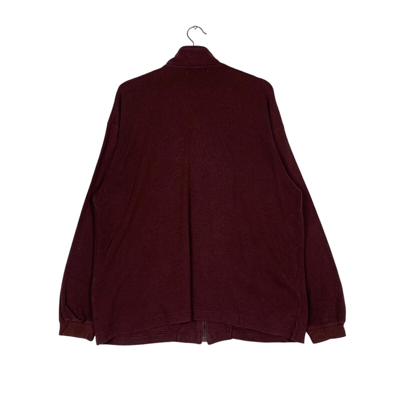 May include: A burgundy-colored long-sleeve zip-up shirt hanging on a hanger. The shirt has a high collar and a full-length zipper. The sleeves have ribbed cuffs. The shirt is made of a soft, textured fabric.