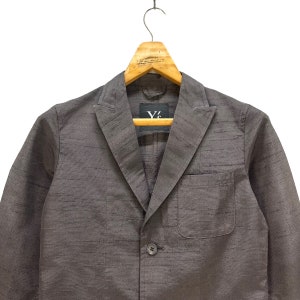 Puede incluir: Una chaqueta gris con un cierre de bot&oacute;n &uacute;nico y un bolsillo en el lado izquierdo. La chaqueta tiene una etiqueta Y's en el interior.