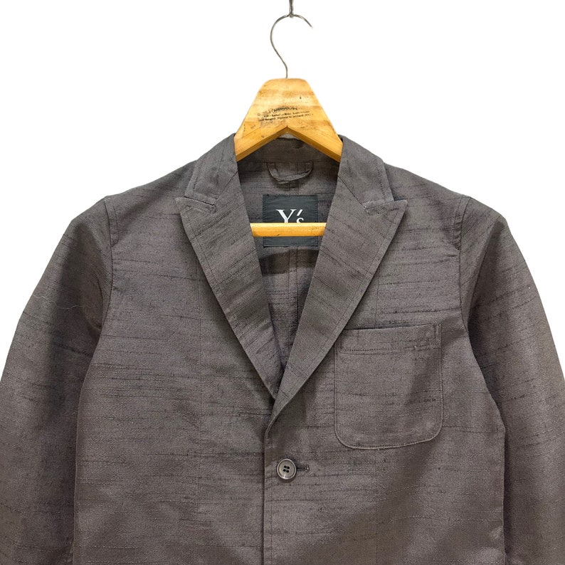 Puede incluir: Una chaqueta gris con un cierre de bot&oacute;n &uacute;nico y un bolsillo en el lado izquierdo. La chaqueta tiene una etiqueta Y's en el interior.