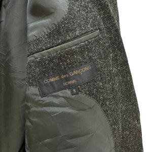 Puede incluir: Una chaqueta de tweed negra y gris con una etiqueta negra que dice "COMME des GAR&Ccedil;ONS HOMME S".