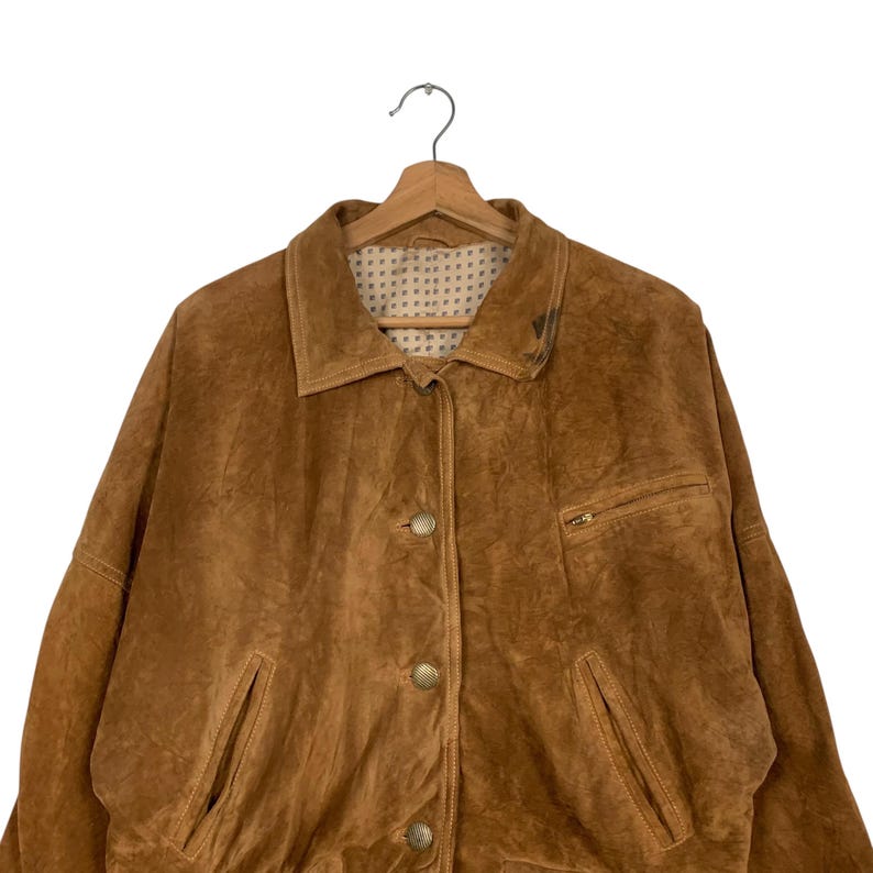 May include: A brown suede jacket with a collared neckline, button-up front, and two front pockets. The jacket is hanging on a wooden hanger. The jacket has a small pocket with a zipper on the right side.