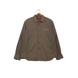Puede incluir: Una camisa abotonada marrón con rayas verticales y dos bolsillos delanteros. La camisa tiene cuello y el texto "URTH STORE'S" está impreso en la parte delantera. La camisa está colgada de una percha de madera.