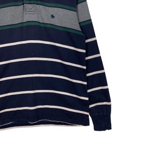 May include: Navy blue long-sleeve shirt with white horizontal stripes. The shirt has a gray and green striped collar and a small embroidered emblem. The sleeves have a ribbed cuff.