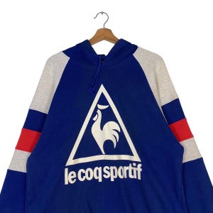 Puede incluir: Una sudadera con capucha azul, gris y roja con un logotipo de gallo blanco y el texto "le coq sportif" en la parte delantera.