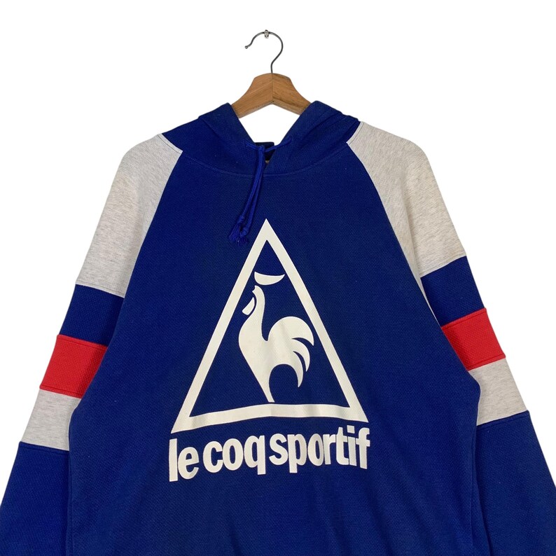 Puede incluir: Una sudadera con capucha azul, gris y roja con un logotipo de gallo blanco y el texto "le coq sportif" en la parte delantera.