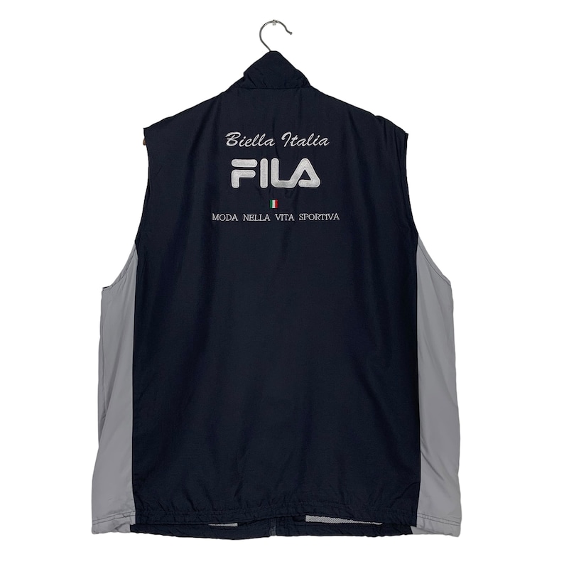 Puede incluir: Un chaleco Fila negro sin mangas con una cremallera blanca y un logotipo Fila blanco en la parte posterior. El chaleco tambi&eacute;n tiene el texto "Biella Italia" y "Moda Nella Vita Sportiva" en blanco. La bandera italiana tambi&eacute;n est&aacute; en la parte posterior del chaleco.