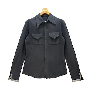 May include: A black long-sleeved shirt with a zipper closure and two flap pockets. The shirt has a collar and the cuffs have the text "NO DEAD ARTISTS" printed on them.