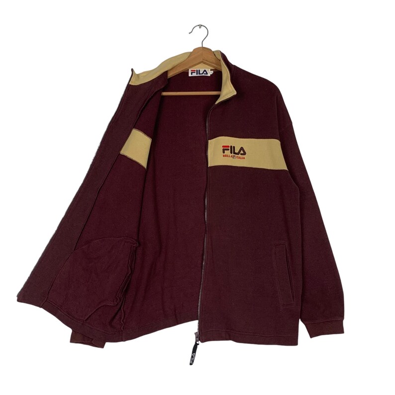 May include: A maroon FILA zip-up jacket with a tan stripe across the chest and collar. The jacket has two pockets and the FILA logo is visible on the chest. The jacket is hanging on a wooden hanger.