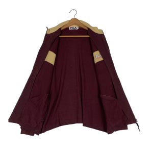 May include: Maroon zip-up jacket with a tan collar and accents. The jacket features two front pockets and the FILA logo on the collar. The jacket is hanging on a hanger.