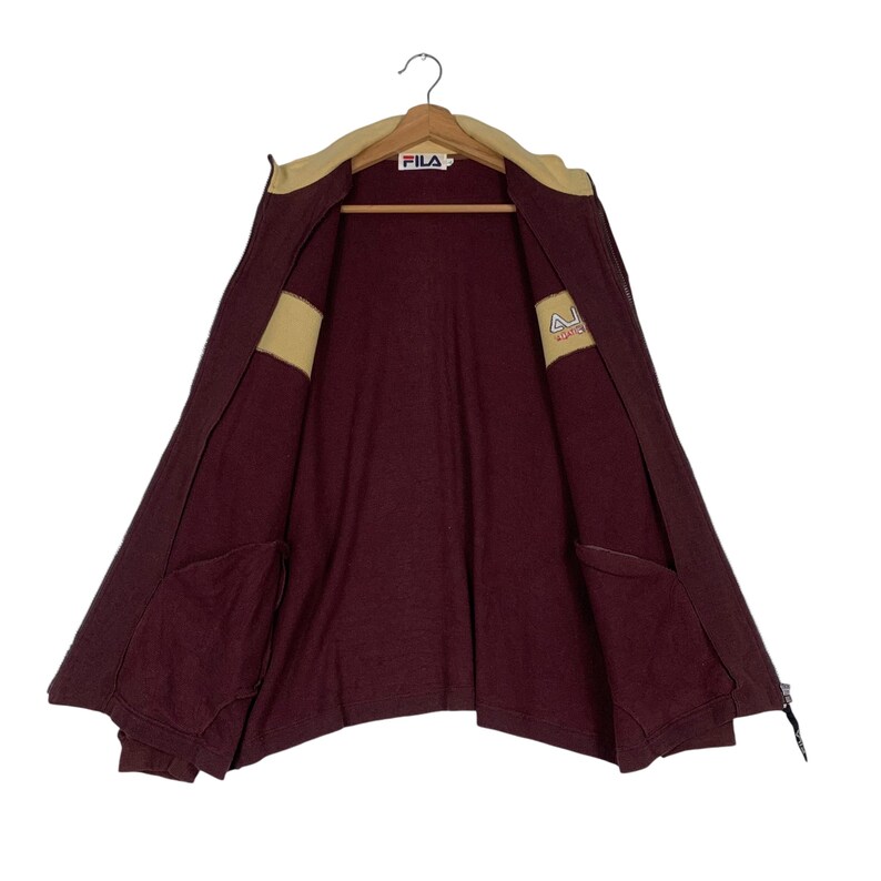 May include: Maroon zip-up jacket with a tan collar and accents. The jacket features two front pockets and the FILA logo on the collar. The jacket is hanging on a hanger.