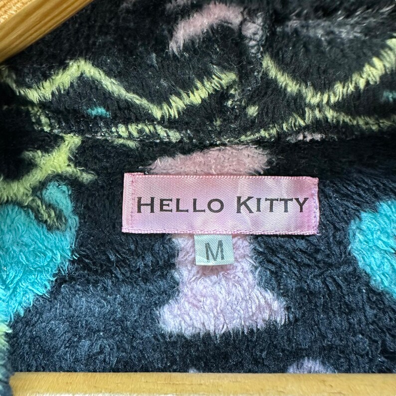 May include: Black fleece jacket with a pink and blue Hello Kitty print. The jacket has a pink label that says "HELLO KITTY" and a size tag that says "M".