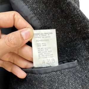 Puede incluir: Una etiqueta de prenda de lana negra con texto blanco que dice "COMME des GARCONS MADE IN JAPAN * FABRIQUE AU JAPON HJ-05050S 100% WOOL 100% LAINE DO NOT BLEACH DO NOT IRON DRY CLEAN ONLY JAVEL INTERDIT NE PAS REPASSER NETTOYAGE A SEC P".