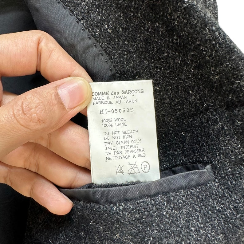 Puede incluir: Una etiqueta de prenda de lana negra con texto blanco que dice "COMME des GARCONS MADE IN JAPAN * FABRIQUE AU JAPON HJ-05050S 100% WOOL 100% LAINE DO NOT BLEACH DO NOT IRON DRY CLEAN ONLY JAVEL INTERDIT NE PAS REPASSER NETTOYAGE A SEC P".