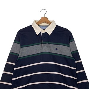 May include: Navy blue long-sleeve rugby shirt with white and grey horizontal stripes. The shirt has a white collar and a small embroidered emblem on the chest. The shirt is hanging on a wooden hanger.