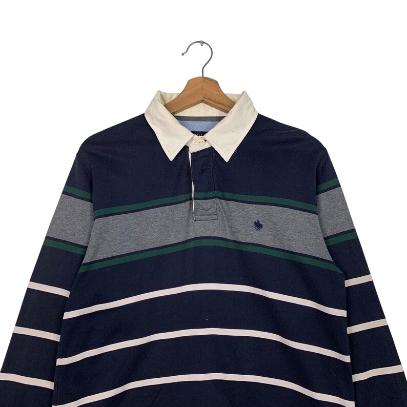 May include: Navy blue long-sleeve rugby shirt with white and grey horizontal stripes. The shirt has a white collar and a small embroidered emblem on the chest. The shirt is hanging on a wooden hanger.