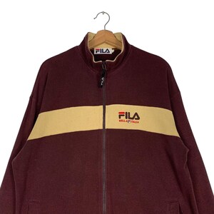 May include: A maroon FILA zip-up jacket with a tan horizontal stripe across the chest. The jacket features the FILA logo on the chest and collar. The jacket is hanging on a wooden hanger.