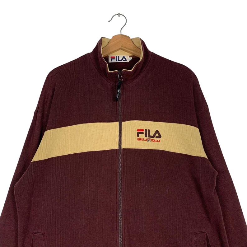 May include: A maroon FILA zip-up jacket with a tan horizontal stripe across the chest. The jacket features the FILA logo on the chest and collar. The jacket is hanging on a wooden hanger.
