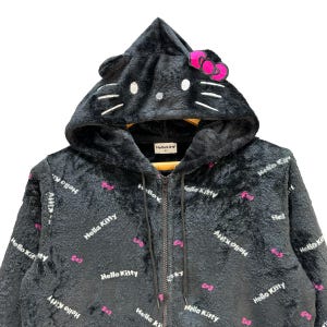 Może przedstawiać: Czarna bluza z kapturem zapinana na zamek z motywem Hello Kitty. Bluza posiada kaptur w kształcie głowy Hello Kitty, z uszami, wąsami i r&oacute;żową kokardą. Materiał pokryty jest powtarzającym się tekstem "Hello Kitty" i grafiką r&oacute;żowych kokardek.