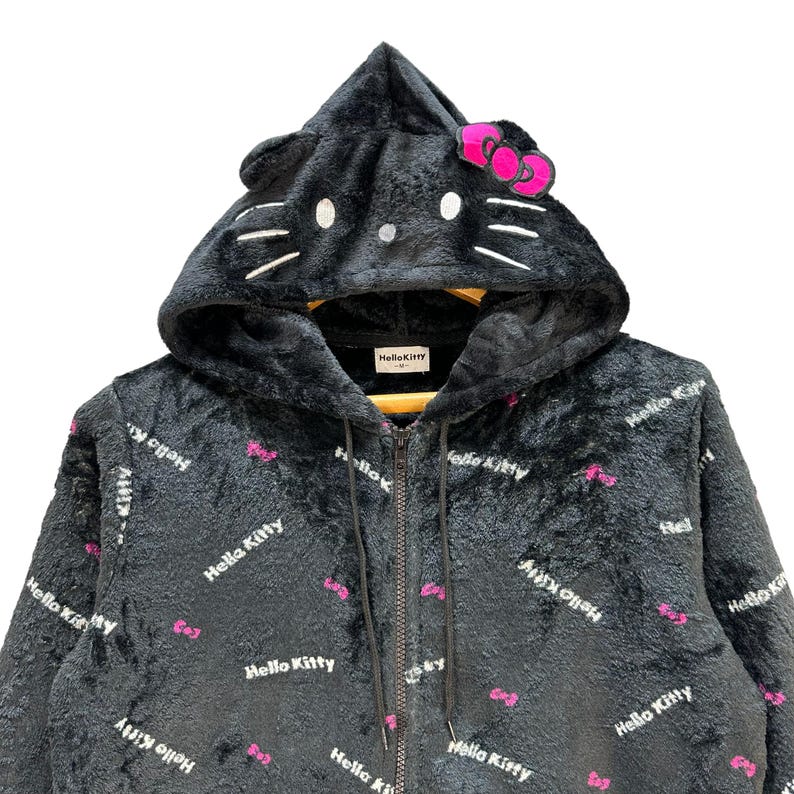 Może przedstawiać: Czarna bluza z kapturem zapinana na zamek z motywem Hello Kitty. Bluza posiada kaptur w kształcie głowy Hello Kitty, z uszami, wąsami i r&oacute;żową kokardą. Materiał pokryty jest powtarzającym się tekstem "Hello Kitty" i grafiką r&oacute;żowych kokardek.