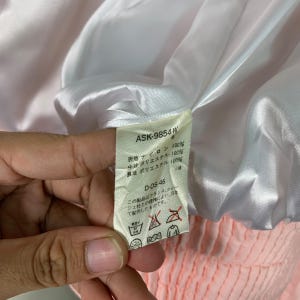 May include: A white tag with black text detailing the fabric composition of a pink jacket. The tag reads "ASK-9854 W 6", "表地 ナイロン 100%", "中 ポリエステル 100%", "裏地 ポリエステル 100%", "D-OS-46", and "この製品はフランス、クレジュ社との提携により、日本て製作したものです."