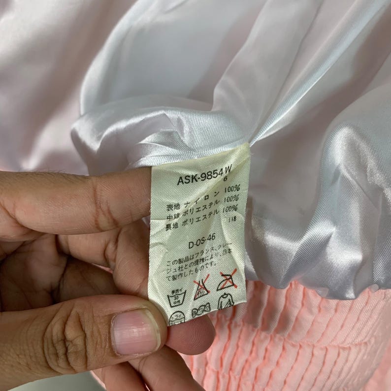 May include: A white tag with black text detailing the fabric composition of a pink jacket. The tag reads "ASK-9854 W 6", "表地 ナイロン 100%", "中 ポリエステル 100%", "裏地 ポリエステル 100%", "D-OS-46", and "この製品はフランス、クレジュ社との提携により、日本て製作したものです."