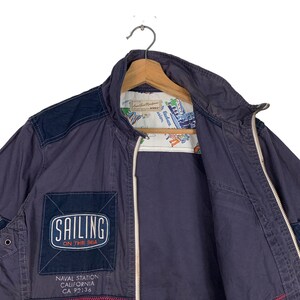 May include: A navy blue jacket with a zip fastening and a patch that says "Sailing on the Sea". The jacket has a navy blue and white patterned lining. The jacket also has a patch that says "Naval Station California CA 92136".