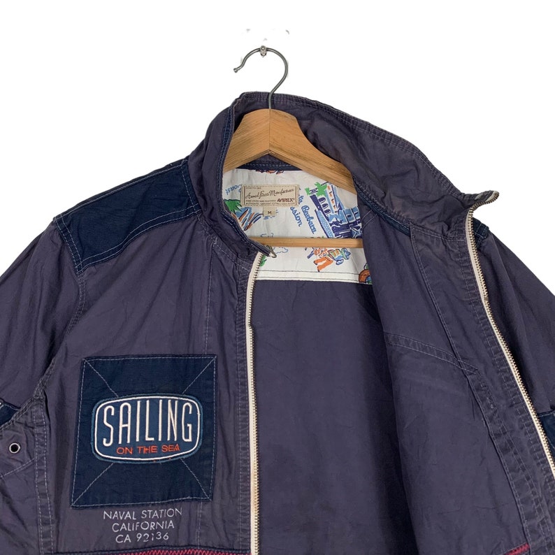 May include: A navy blue jacket with a zip fastening and a patch that says "Sailing on the Sea". The jacket has a navy blue and white patterned lining. The jacket also has a patch that says "Naval Station California CA 92136".