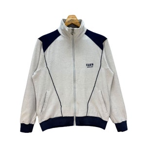 Può includere: Una giacca da tuta con zip grigia e blu navy con il testo "CLUB adidas" ricamato sul davanti.