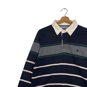 May include: A long-sleeved rugby shirt with a navy blue and white striped pattern. The shirt features gray and green horizontal stripes across the chest, a white collar, and a small embroidered logo. The shirt is hanging on a wooden hanger.