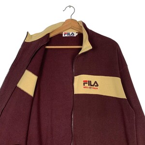May include: A maroon zip-up jacket with a cream-colored collar and stripe across the chest. The jacket features the Fila logo on the chest and collar. The jacket is hanging on a wooden hanger.