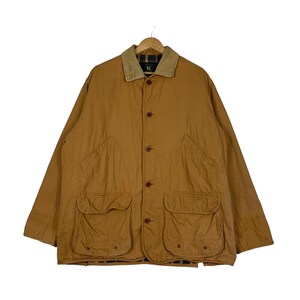 May include: A tan-colored jacket with a corduroy collar and four front pockets. The jacket has a button-up closure and is hanging on a wooden hanger. The jacket appears to be made of a durable, possibly water-resistant material.