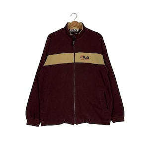 May include: A maroon zip-up jacket with a tan horizontal stripe across the chest. The jacket features the brand name "FILA" in white on the chest and collar. The jacket is hanging on a metal hanger.