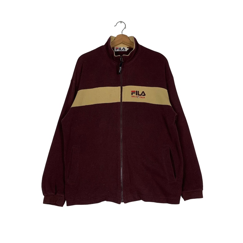 May include: A maroon zip-up jacket with a tan horizontal stripe across the chest. The jacket features the brand name "FILA" in white on the chest and collar. The jacket is hanging on a metal hanger.