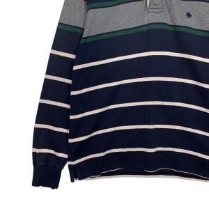 May include: A long-sleeved polo shirt with horizontal stripes in navy blue and white. The top section features gray and green stripes. The shirt has a classic collar and a small embroidered logo on the chest.