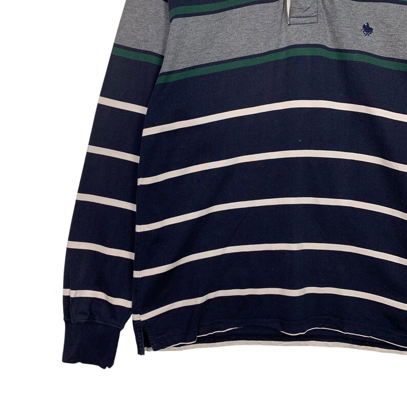 May include: A long-sleeved polo shirt with horizontal stripes in navy blue and white. The top section features gray and green stripes. The shirt has a classic collar and a small embroidered logo on the chest.