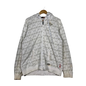 May include: White zip-up hoodie with a full-coverage all-over print of a logo design. The hoodie has a drawstring hood and a zipper closure.