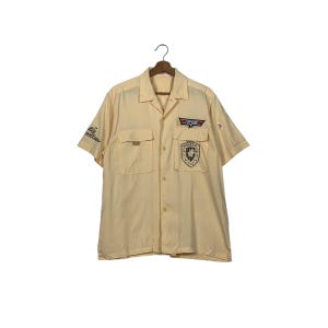 Può includere: Una camicia gialla a maniche corte con bottoni, con una toppa sul lato destro che dice "Top Gun" e un'altra toppa sul lato sinistro che dice "Smiles".