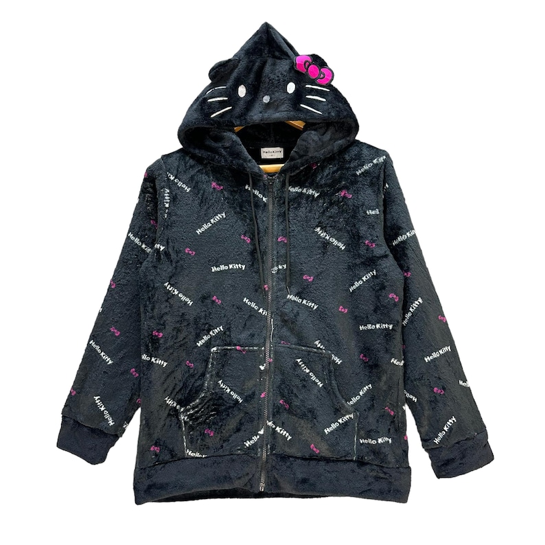 Może przedstawiać: Czarna bluza z kapturem zapinana na zamek z motywem Hello Kitty. Bluza posiada kaptur w kształcie głowy Hello Kitty, z uszami i r&oacute;żową kokardą. Materiał pokryty jest powtarzającym się tekstem "Hello Kitty" i r&oacute;żowymi akcentami kokard.