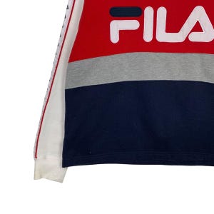May include: A color-blocked sweatshirt featuring the brand name "FILA" in white block letters. The top section is red, followed by a gray band, and a navy blue lower section. The left sleeve has a white base with a red and blue stripe.