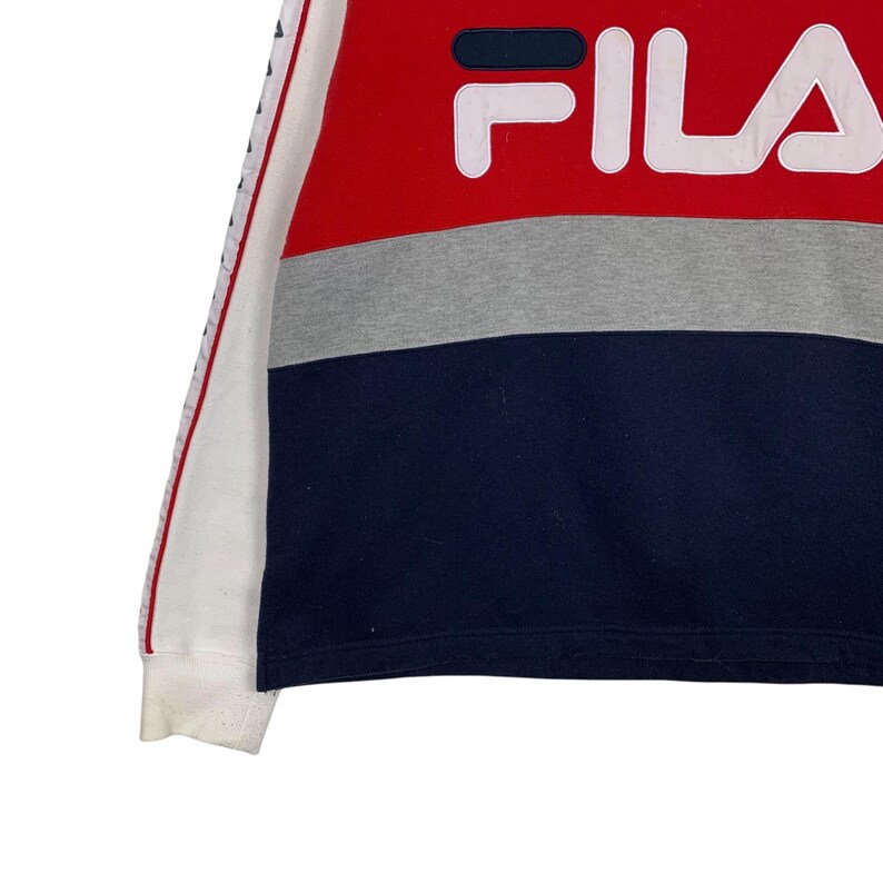 May include: A color-blocked sweatshirt featuring the brand name "FILA" in white block letters. The top section is red, followed by a gray band, and a navy blue lower section. The left sleeve has a white base with a red and blue stripe.